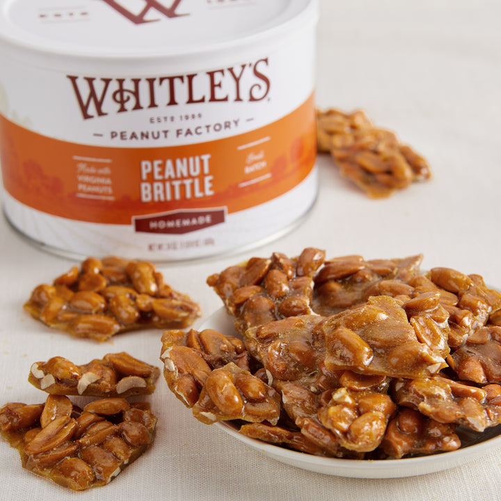 Dish filled with Whitley's Peanut Brittle surrounded by loose pieces with tin in the background on linen tablecloth