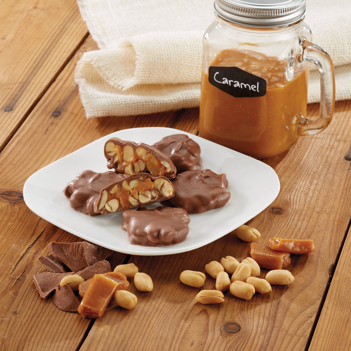 Dish filled with Milk Chocolatey Delights with jar of caramel, loose milk chocolate, caramel pieces, and penuts on wood table.